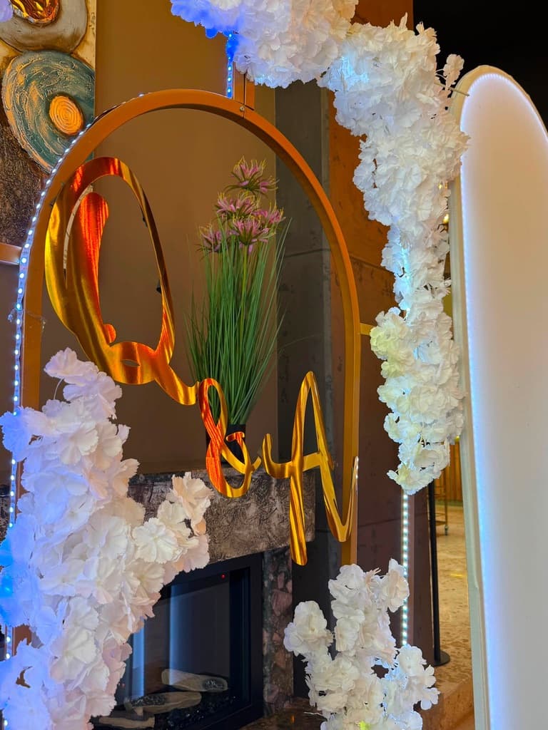 Detaj i një harku të artë me lule të bardha dhe iniciale të personalizuara
